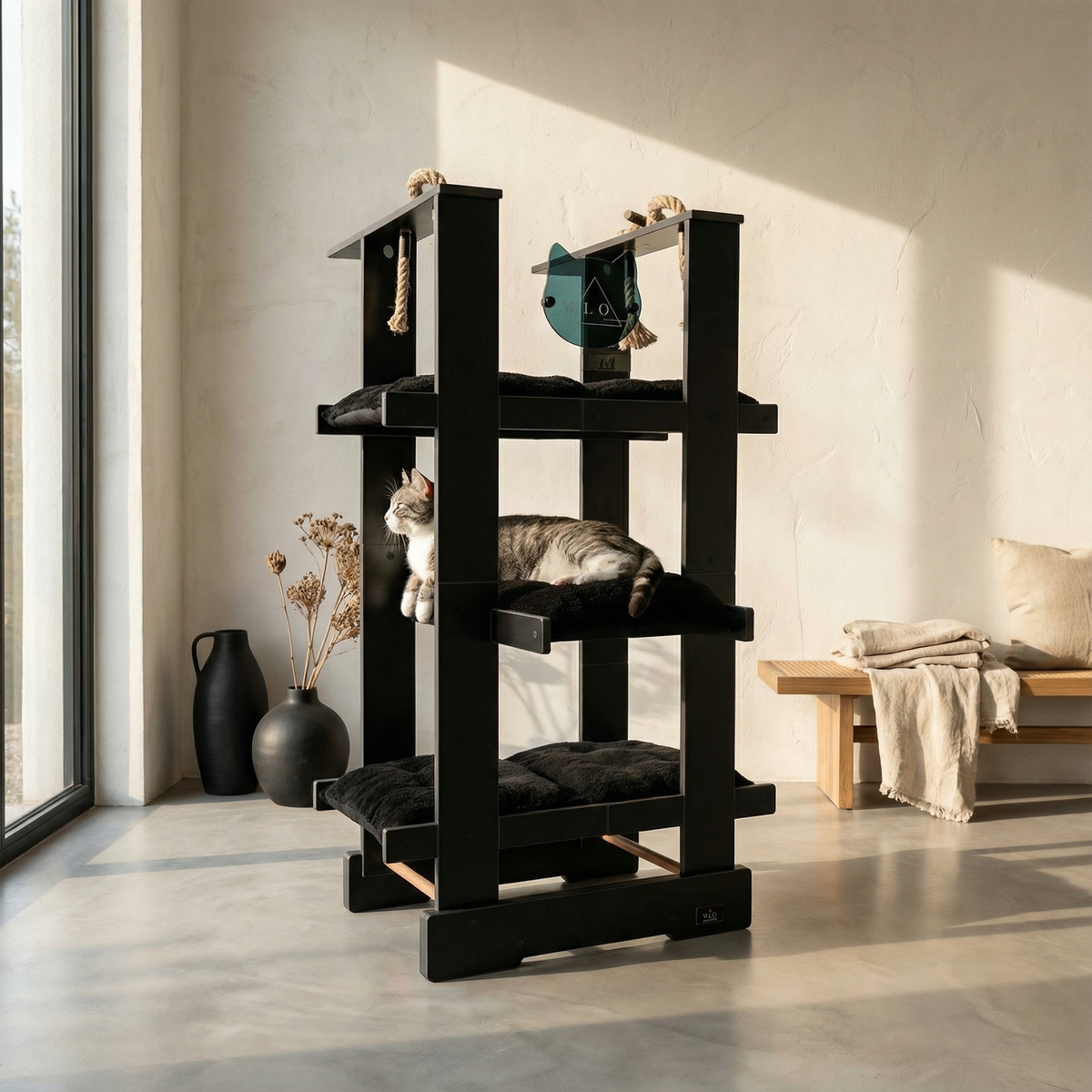 Black cat tree with a cat sitting on it in a room with a window and bench.